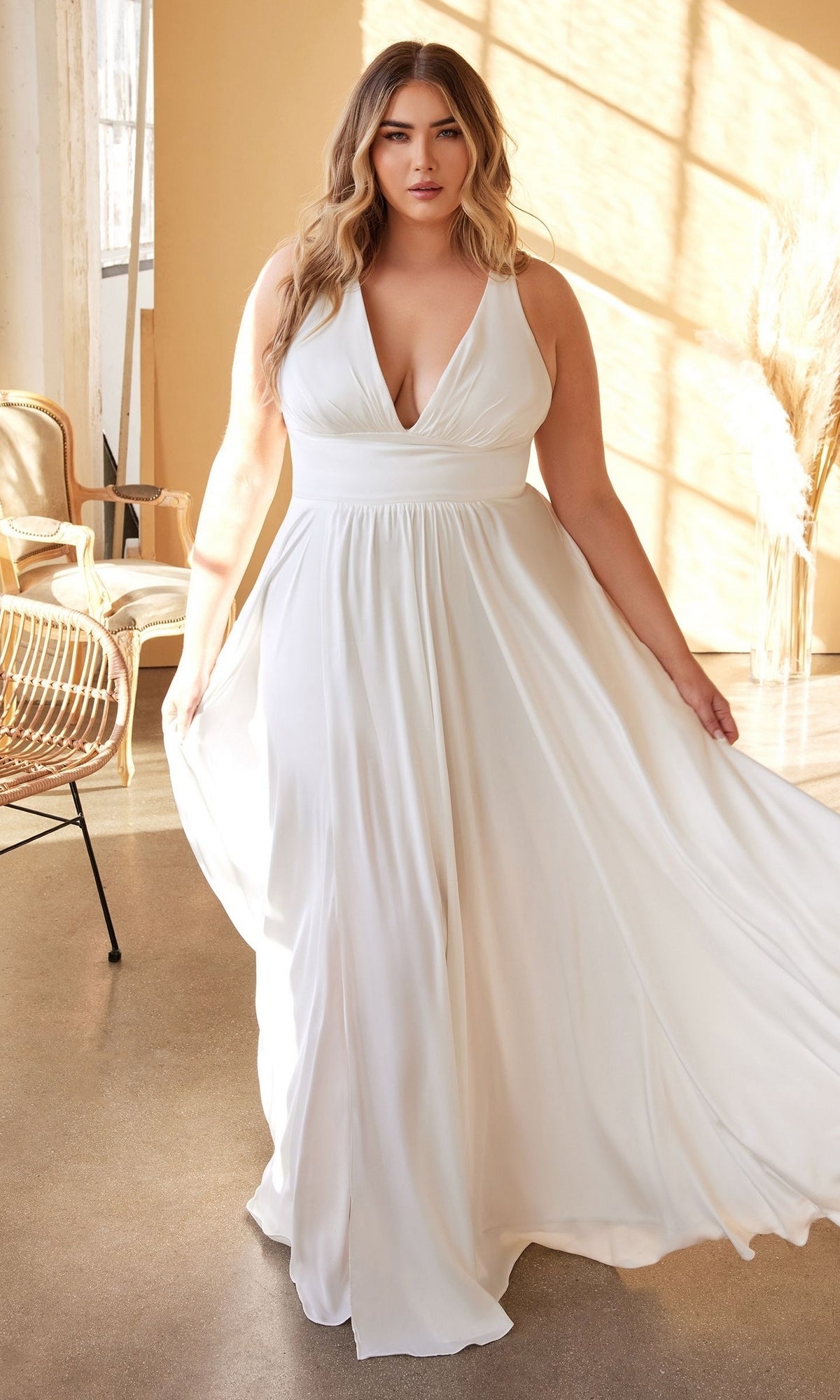 A woman stands indoors in the Long Plus-Size White Dress: Ladivine 7469WW, a sleeveless, floor-length off white gown. She gently holds out the skirt as natural light streams through large windows behind her.
